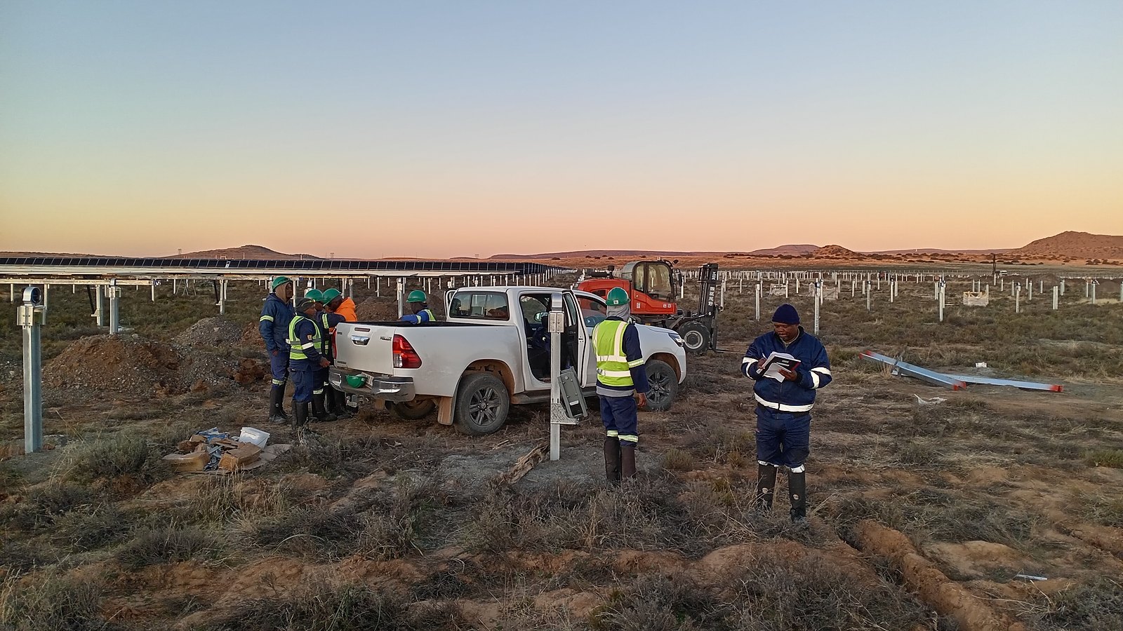 A utility-scale solar farm under construction at dusk: completed rows of panels on one side, driven piles awaiting racking on the other, with the crew and a telehandler on site.